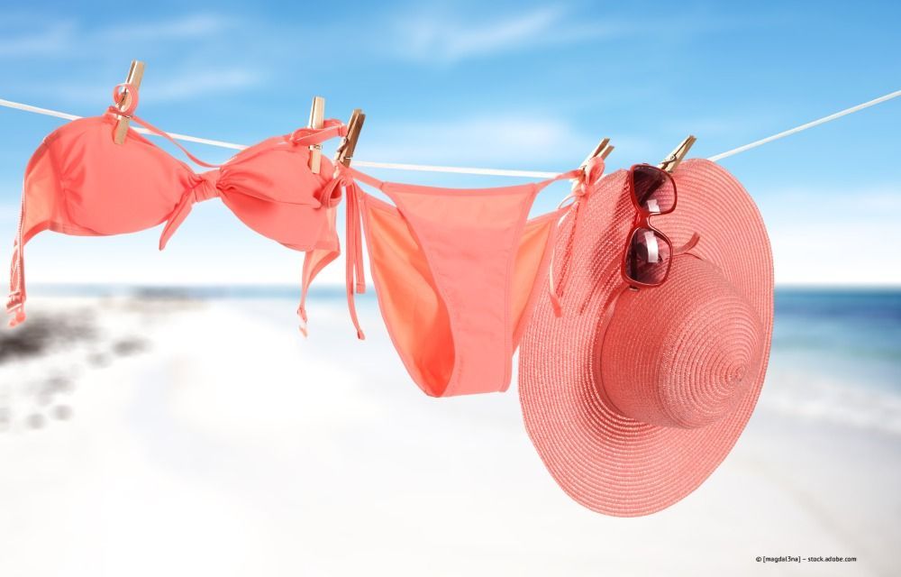 Ein pinkfarbener Bikini, ein Hut und eine Sonnenbrille hängen an einer Wäscheleine am Strand.