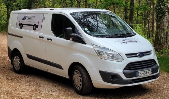 Une camionnette Ford Transit blanche est garée sur un sentier boisé, avec un dessin de camionnette sur son panneau latéral.