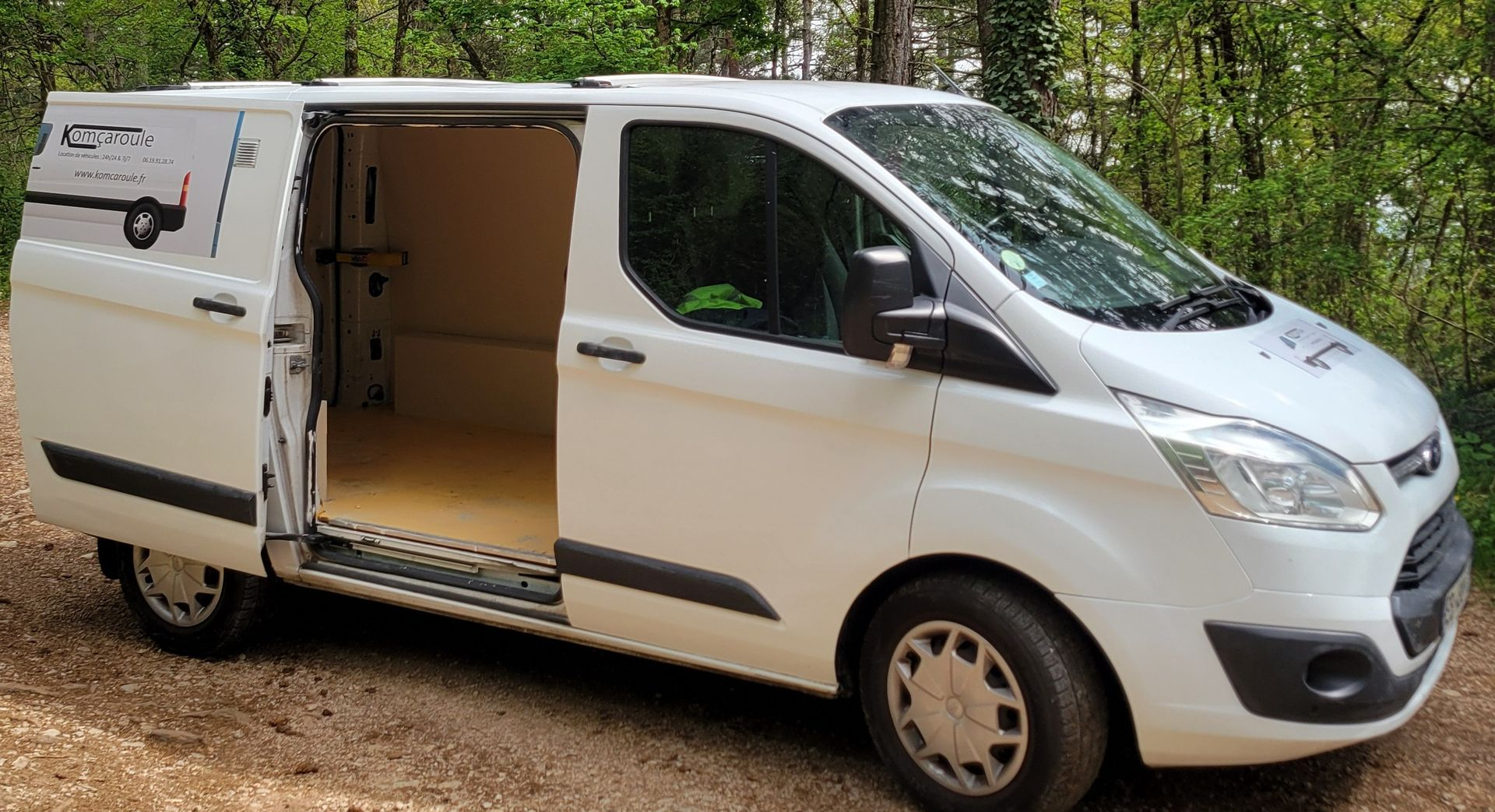Une camionnette blanche, porte latérale ouverte, garée sur un chemin de terre dans une zone boisée.