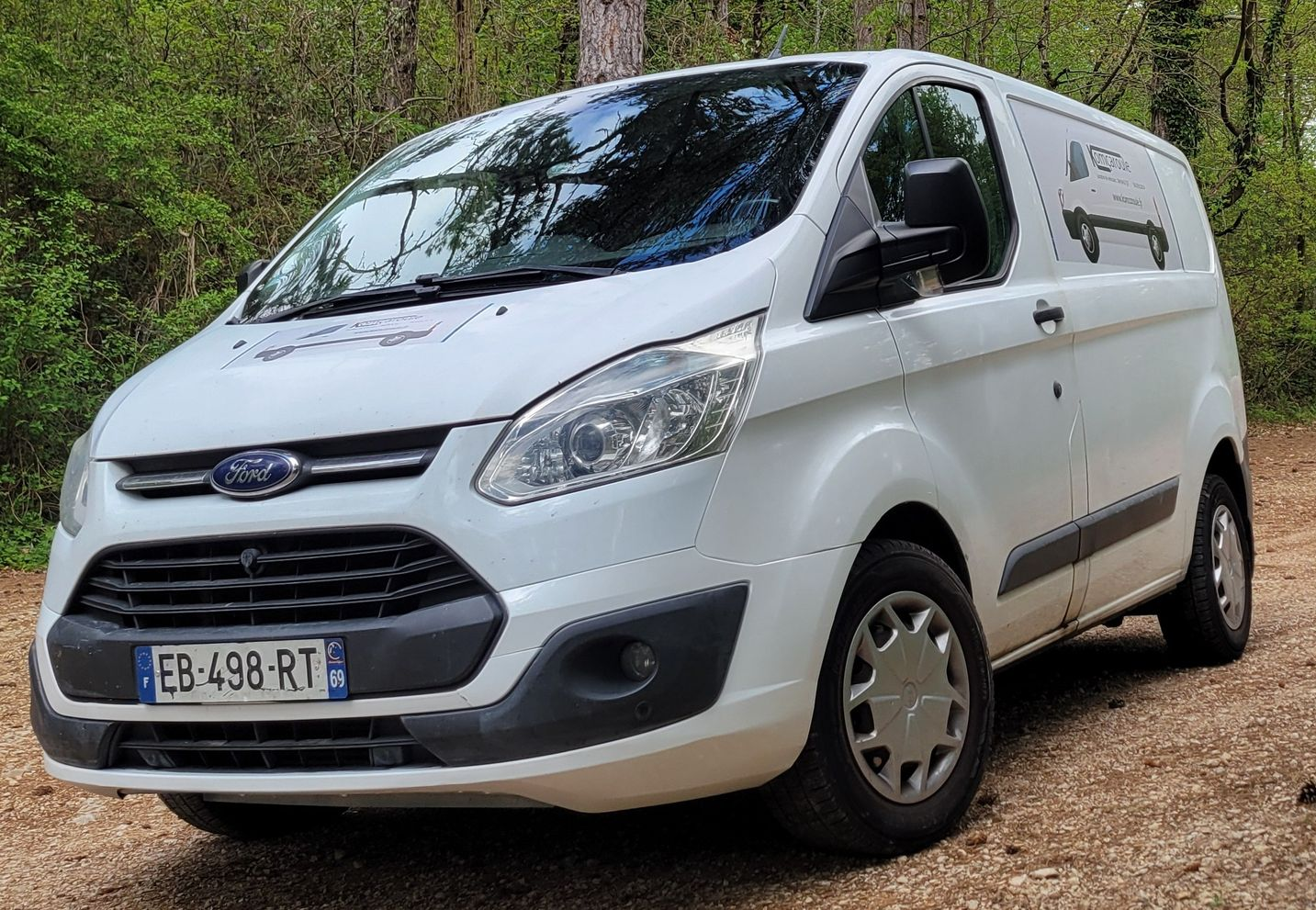 Fourgonnette Ford Transit Custom blanche garée sur un chemin de terre dans une zone boisée.