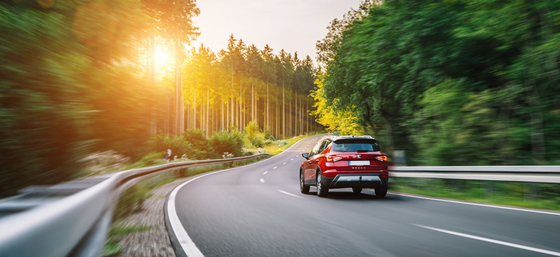 Une voiture rouge roule sur une route sinueuse à travers une forêt verdoyante au coucher du soleil.