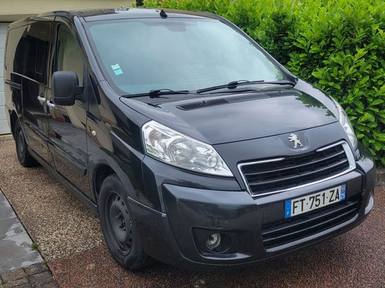 Une camionnette Peugeot Expert noire est garée sur une allée de gravier, devant une haie verte.