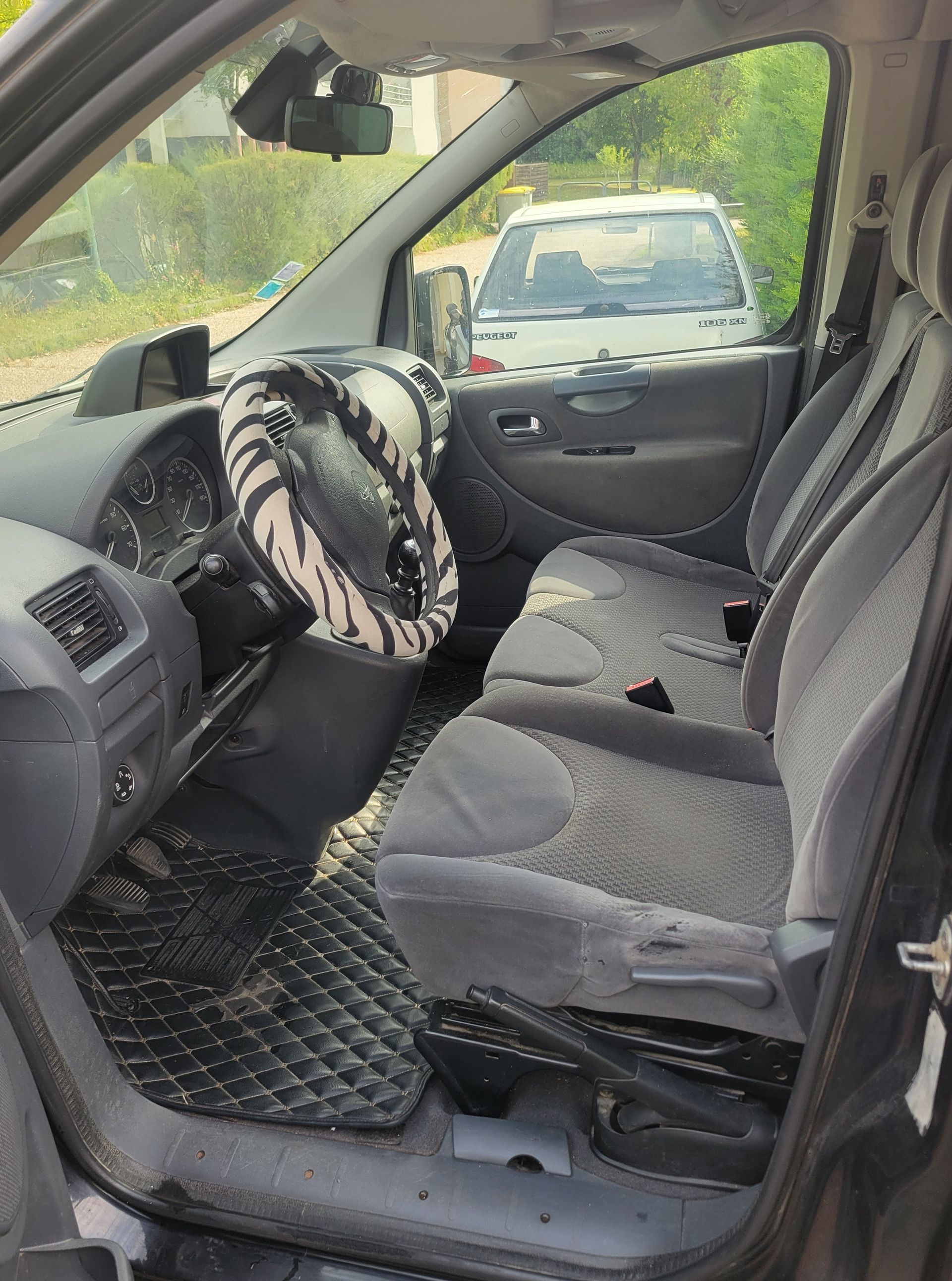 Intérieur d'un véhicule avec des sièges en tissu gris, couvre-volant à imprimé tigre et tapis de sol noirs texturés.