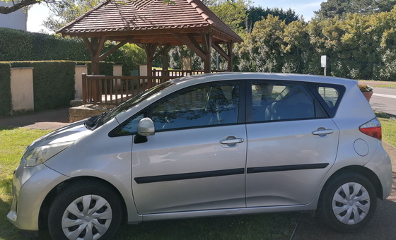Une voiture à hayon argentée garée sur une pelouse devant un kiosque en bois par une journée ensoleillée.