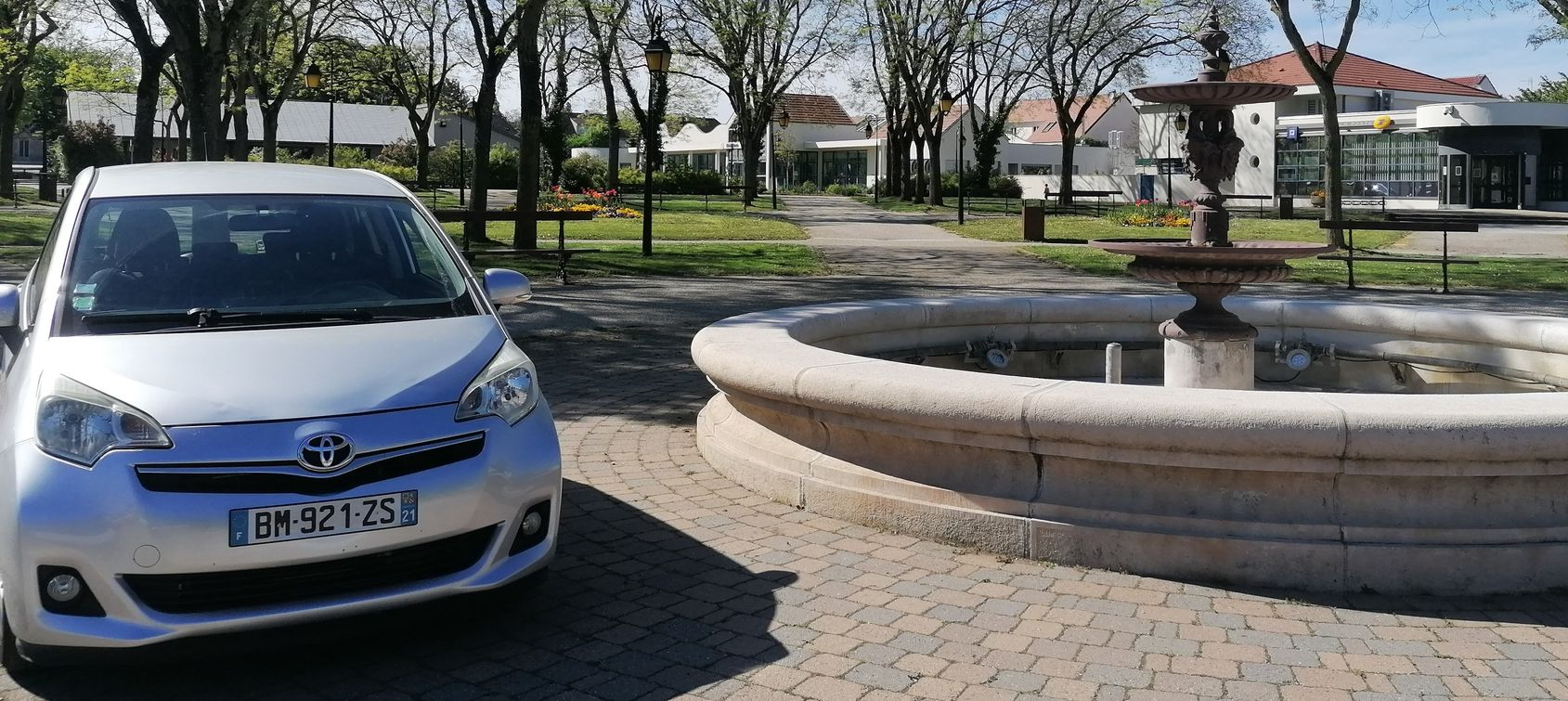 Une Toyota argentée garée à côté d'une fontaine en pierre dans un parc ensoleillé, avec des arbres et des bâtiments en arrière-plan.