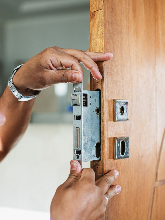 Des mains installent une serrure métallique dans un cadre de porte en bois.
