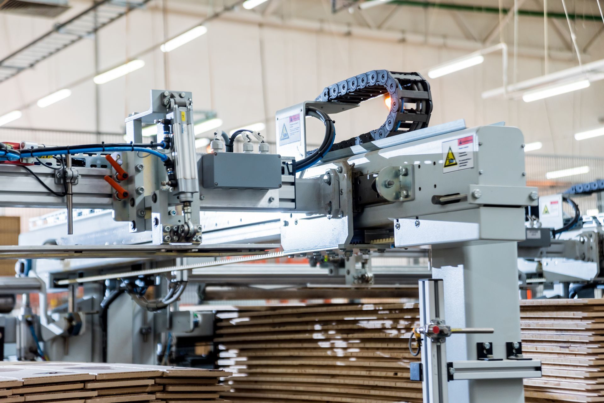 Industriemaschine mit Roboterarm und Kabelträger über Stapeln von Kartonbögen in einer Fabrikumgebung.