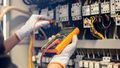 Eine Person mit Arbeitshandschuhen testet mit einem digitalen Multimeter elektrische Bauteile im Inneren eines industriellen Schaltschranks.