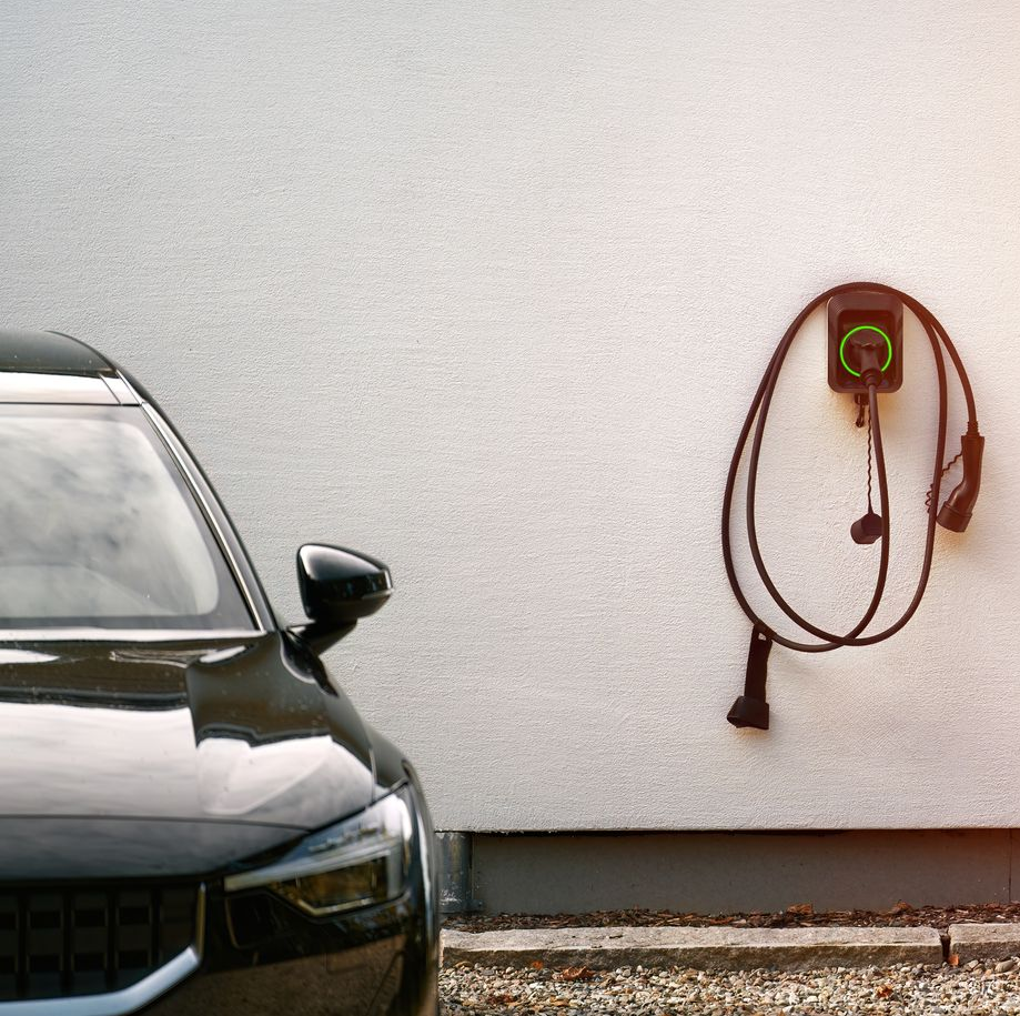 Ein schwarzes Elektrofahrzeug parkt neben einer weißen Wand, an der eine Wallbox-Ladestation mit einem aufgerollten Kabel montiert ist.