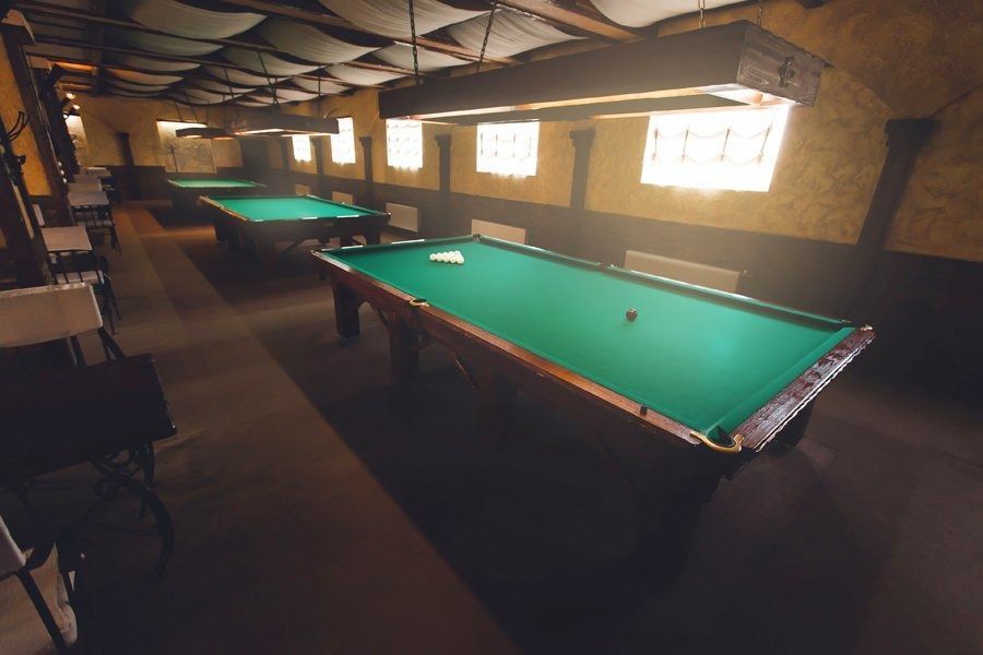 Una sala de billar poco iluminada, con varias mesas de billar revestidas de fieltro verde, paredes de madera e iluminación cenital.
