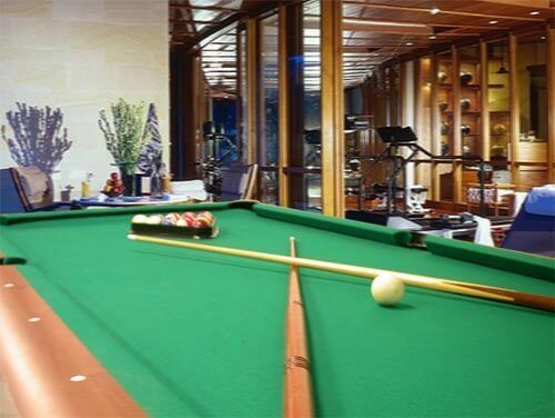 Una mesa de billar verde con bolas y tacos en una sala de lujo, con una zona de fitness al fondo.