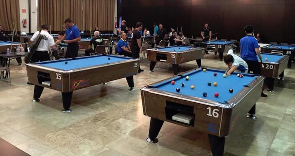 Torneo de billar en un gran salón. Varias mesas de billar marrones están en uso y los jugadores están en acción.