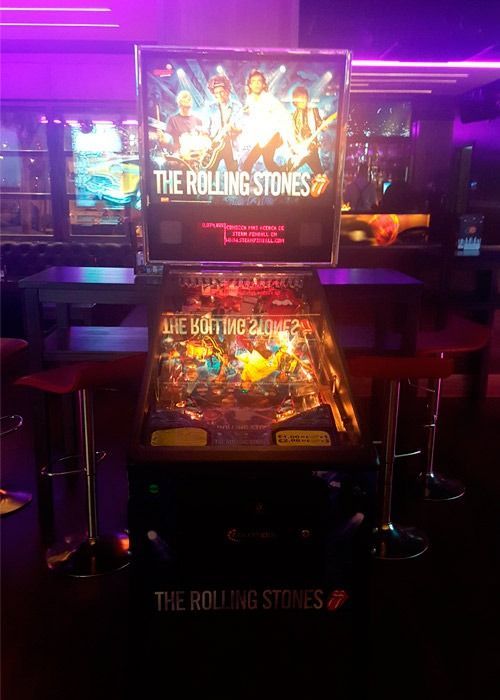 Una máquina de pinball de los Rolling Stones en un bar con poca luz y detalles morados. El cristal trasero luce una foto de la banda.