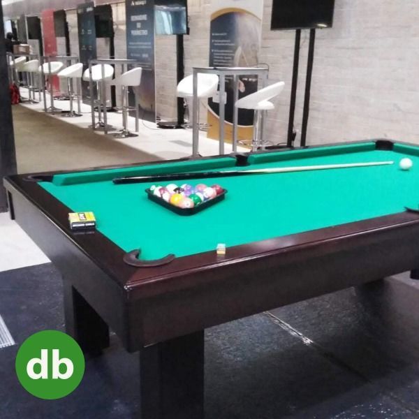 Una mesa de billar con un soporte de bolas, un taco y una bola sobre el tapete verde. El entorno es un bar o sala de juegos.