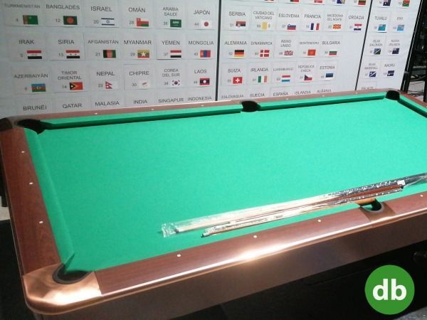 Una mesa de billar verde con detalles marrones, un soporte para el taco y banderas en el fondo.