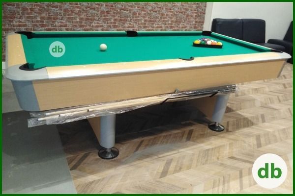 Una mesa de billar con fieltro verde, una bola blanca y un soporte de bolas, lista para jugar. La mesa tiene un marco color canela y se apoya sobre un suelo de madera.