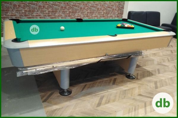 Mesa de billar con fieltro verde, bolas colocadas y una bola blanca. La mesa tiene un marco claro y está en una habitación con una pared de ladrillo.