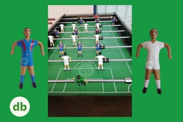 Mesa de futbolín con jugadores con uniformes azules y blancos, figuras de jugadores con uniformes azules y blancos a cada lado.