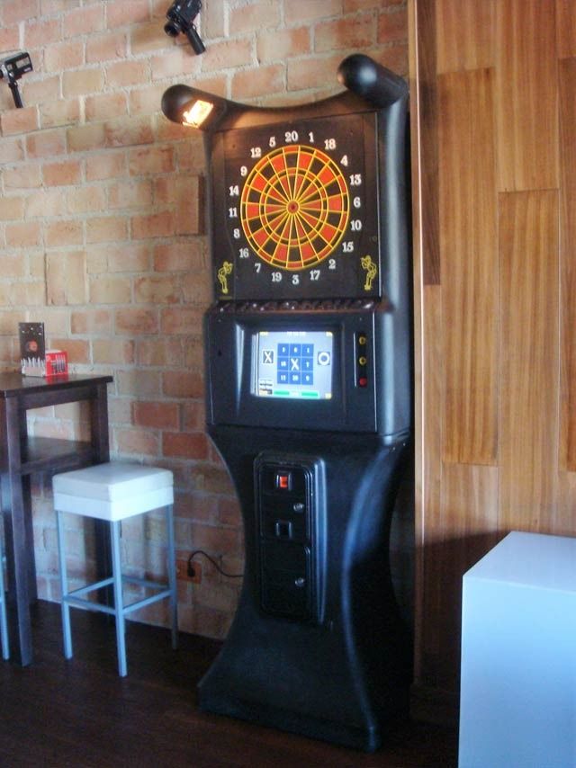 Máquina de dardos con pantalla digital, mueble negro, colocada contra una pared de ladrillos y revestimiento de madera, con un taburete y una mesa cerca.