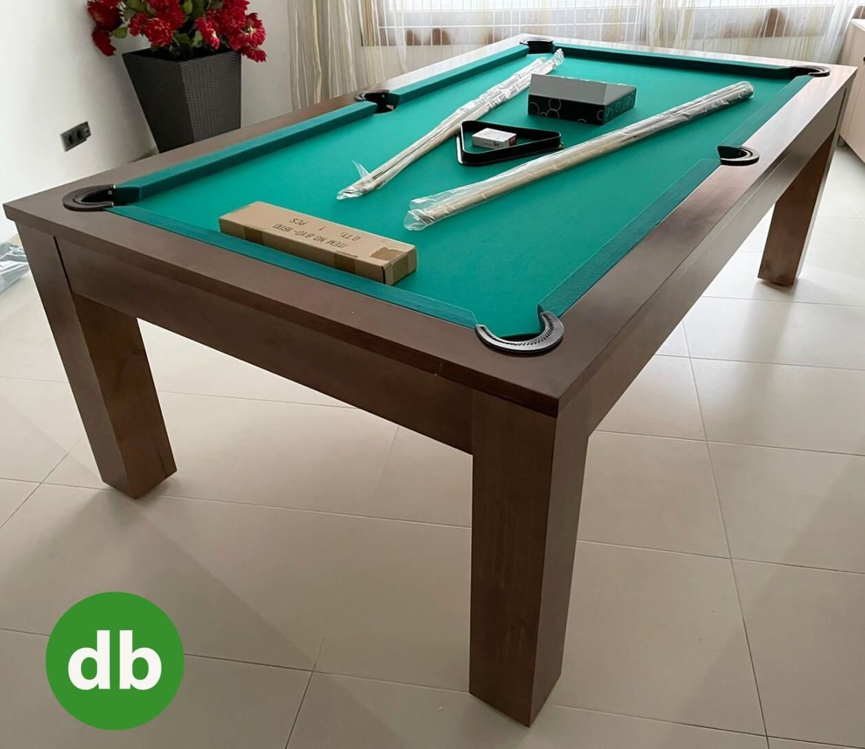 Una mesa de billar con marco de madera y fieltro verde. Sobre ella hay dos tacos y otros accesorios.