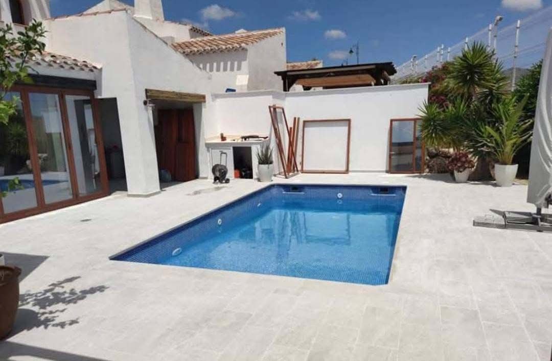 Patio trasero con piscina rectangular, terraza blanca y casa de color claro bajo un cielo soleado.