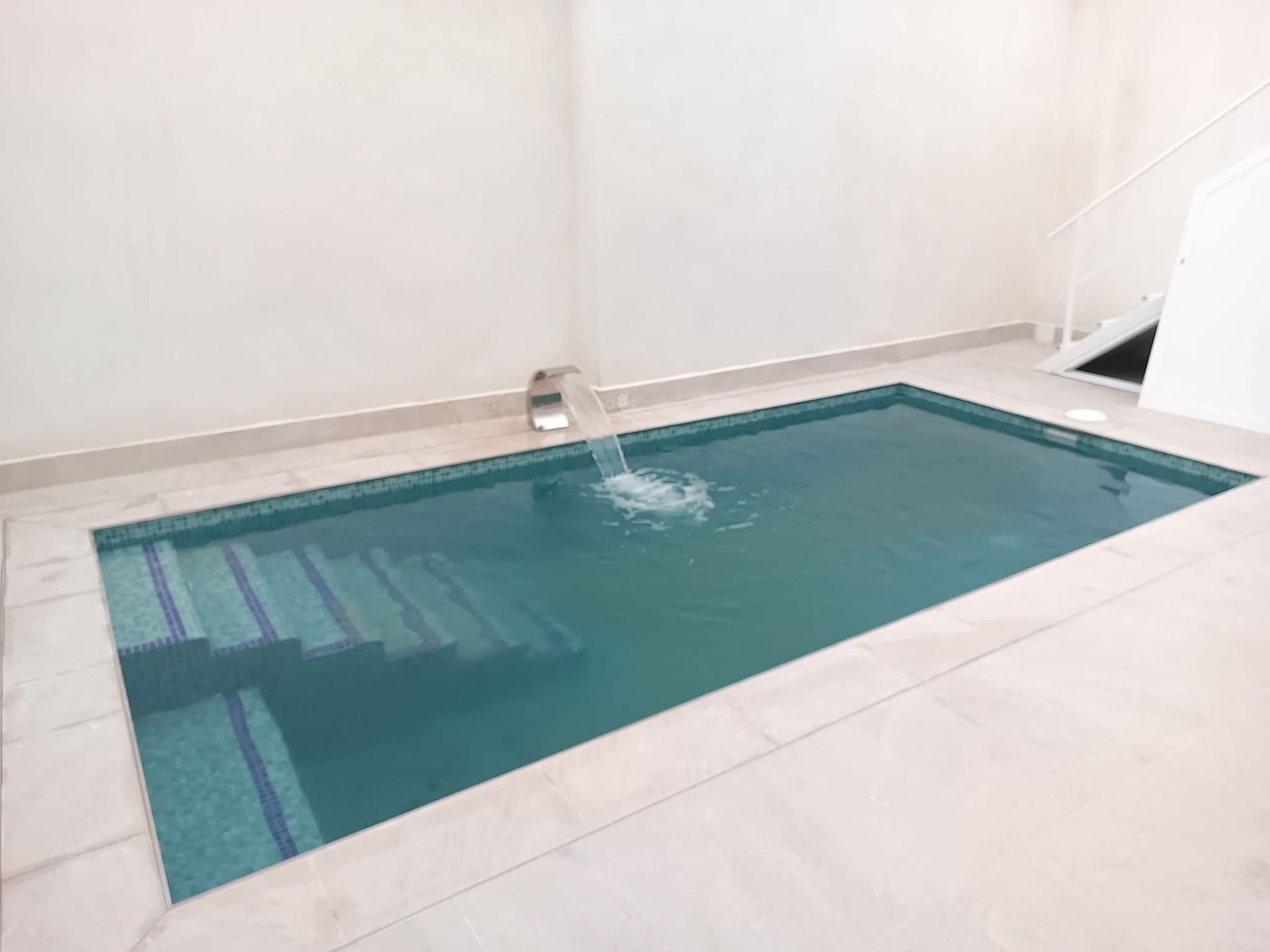 Pequeña piscina rectangular cubierta con escalones, cascada y azulejos azul claro.