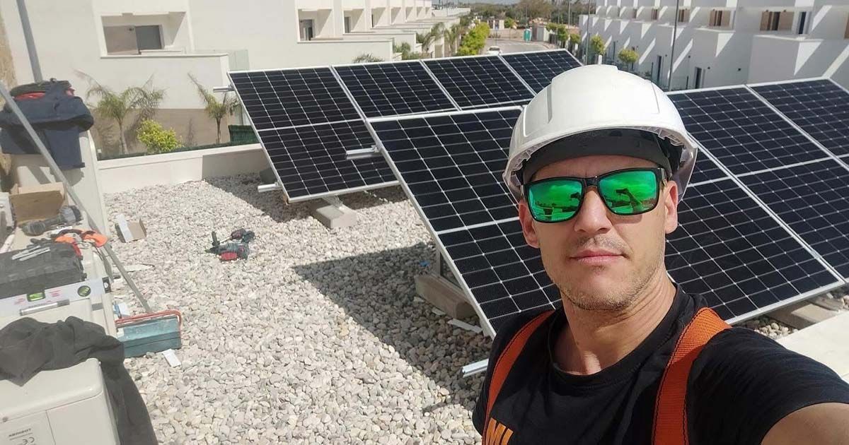 Hombre con casco y gafas de sol, selfie con paneles solares en el tejado a plena luz del sol.
