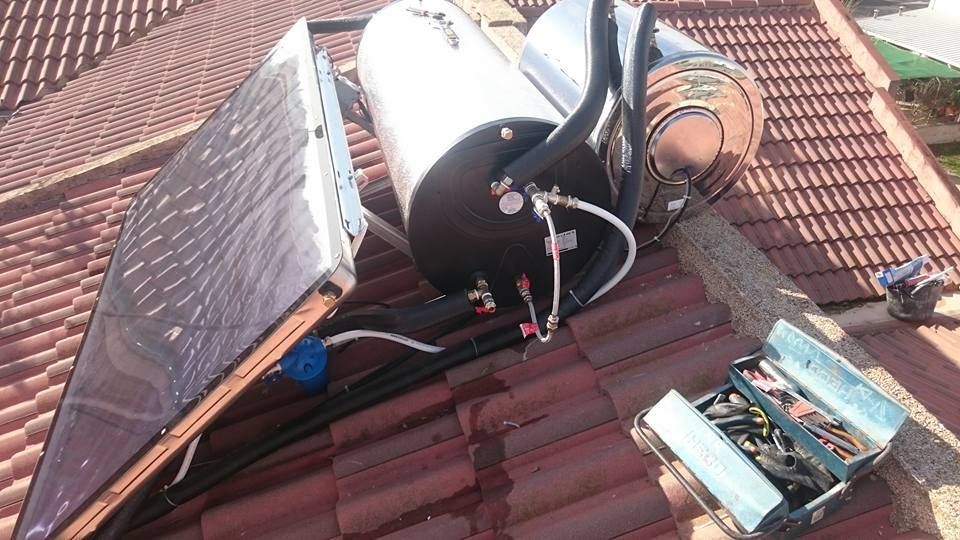 Sistema de calentador solar de agua sobre un tejado de tejas rojas, con depósito de aguas negras, panel solar y caja de herramientas.