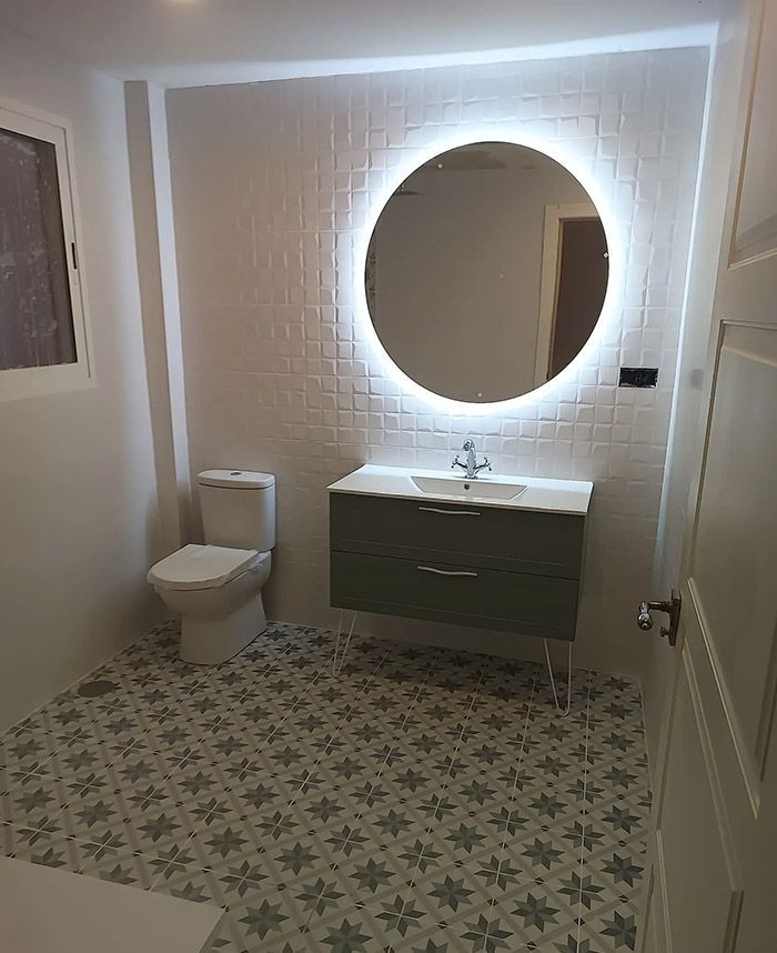 Baño con pared de azulejos blancos, gran espejo redondo con luz, tocador verde y baldosas estampadas en el suelo.