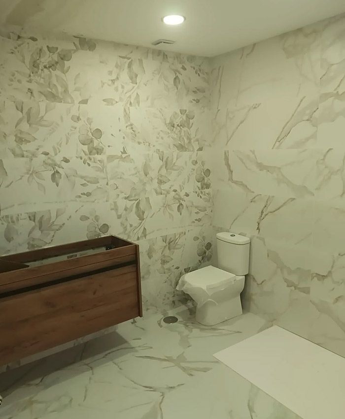 Baño con azulejos de pared con aspecto de mármol y motivos florales, tocador de madera e inodoro, con luz de techo brillante.
