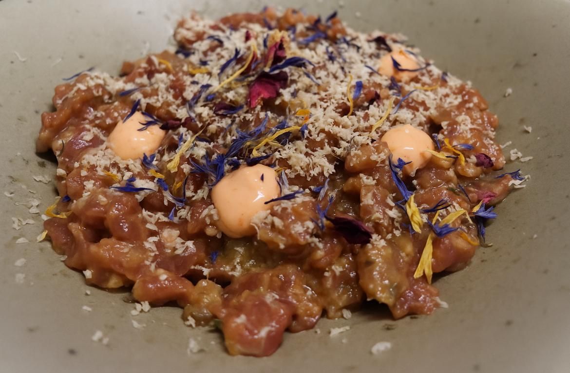 Plato de carne cortada en cubitos con cobertura de migas, cucharadas y pétalos de flores azules en un recipiente gris.