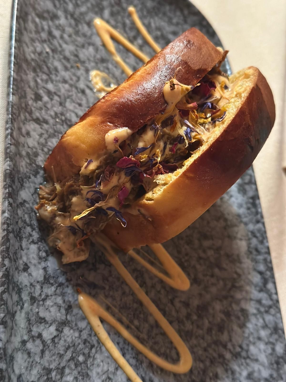 Sándwich abierto sobre plato marmolado, cubierto con flores de colores y un chorrito de salsa.