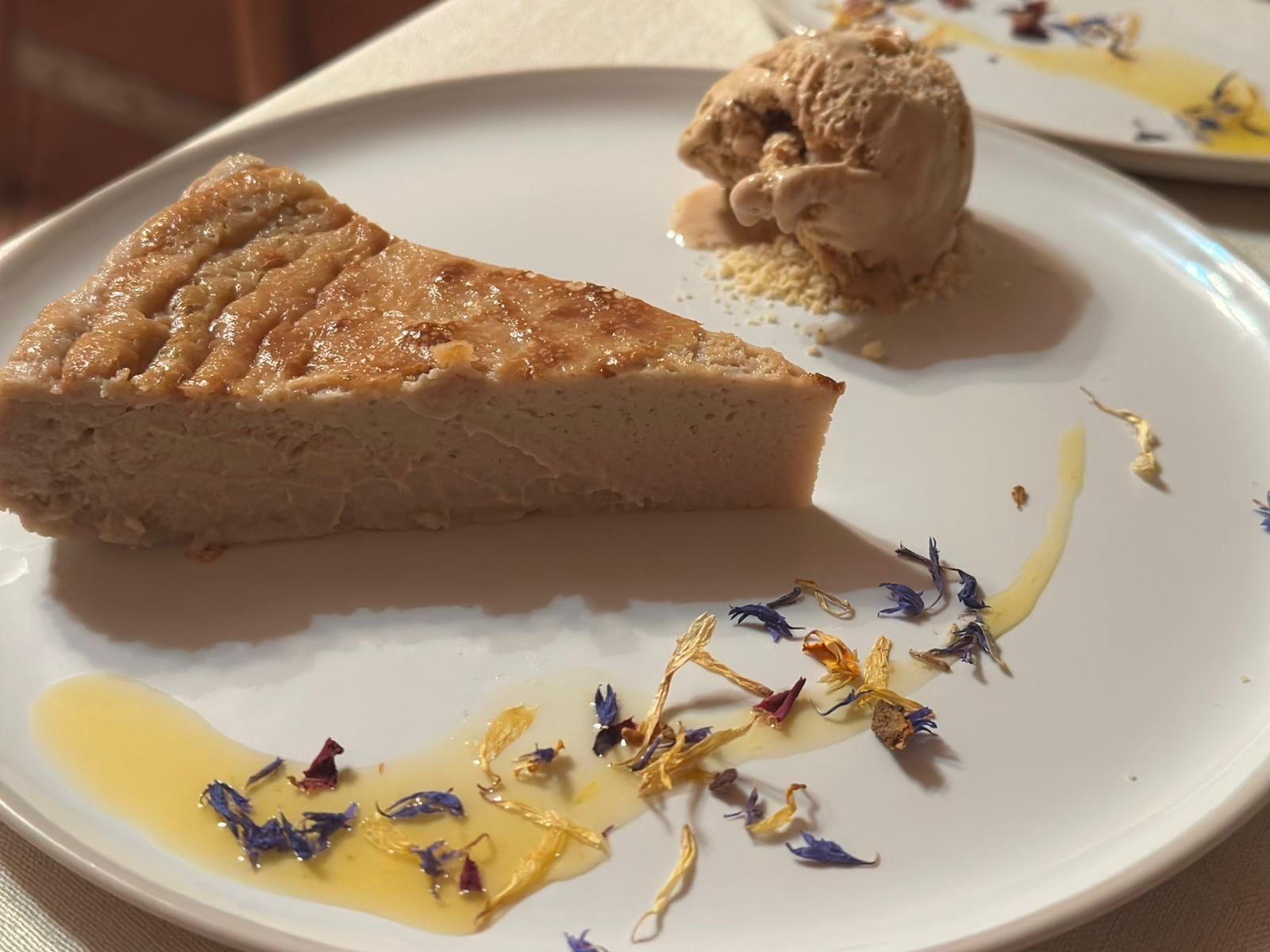 Rebanada de pastel con helado, llovizna y pétalos de flores en un plato blanco.