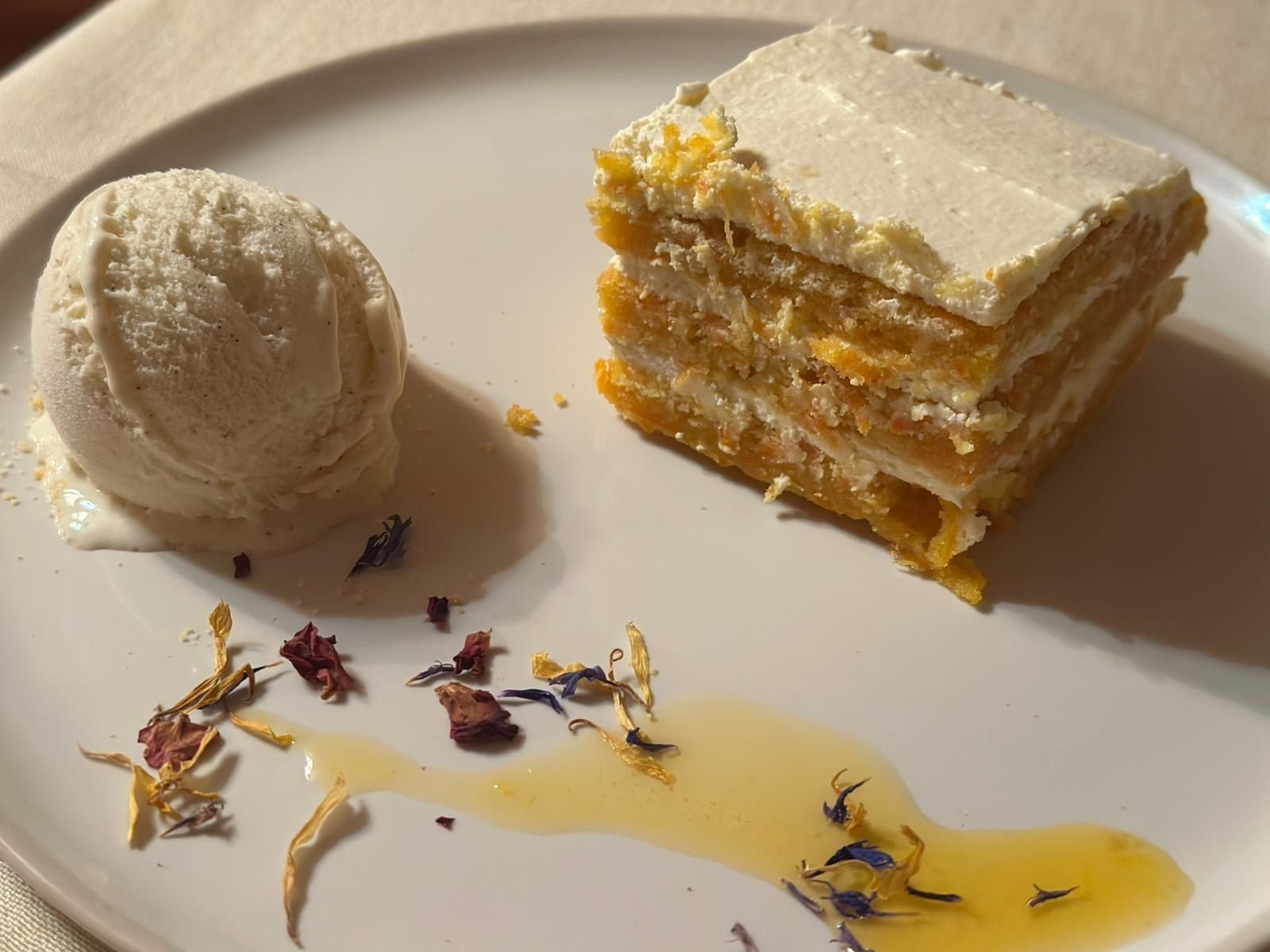 Pastel amarillo de capas con glaseado blanco, bola de helado y chorrito de miel sobre un plato blanco.