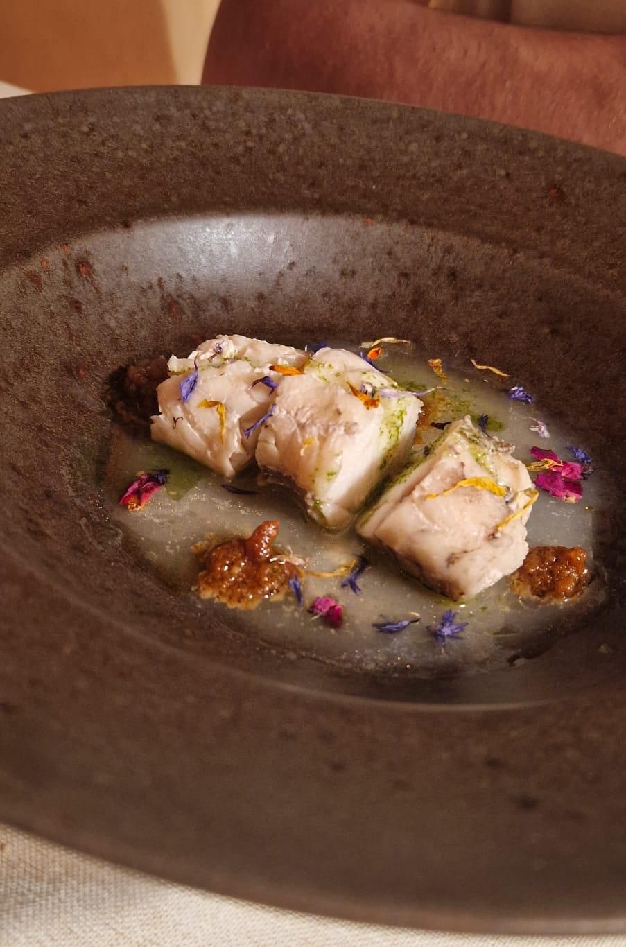 Un plato de pescado de color claro en caldo, adornado con flores y servido en un recipiente oscuro.