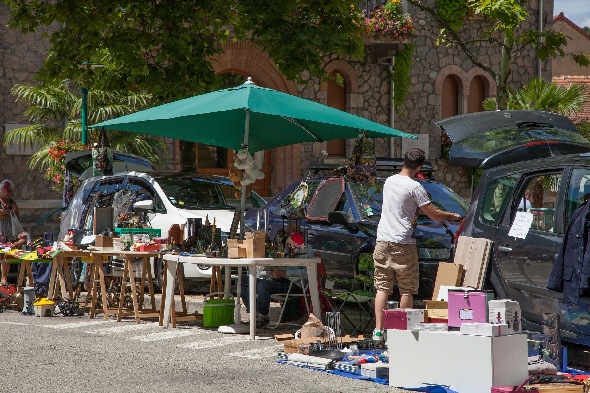 Une personne se tient devant un étal d'un marché aux puces en plein air, sous un grand parasol, à côté de voitures aux coffres ouverts.
