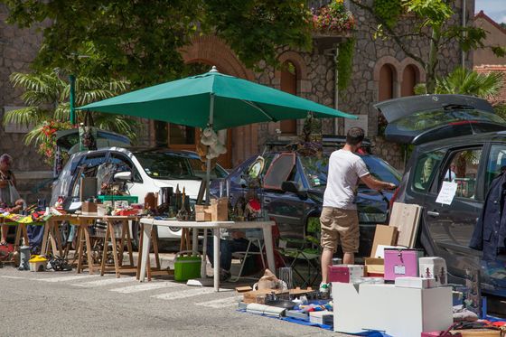 Une personne se tient devant un étal d'un marché aux puces en plein air, sous un grand parasol, à côté de voitures aux coffres ouverts.