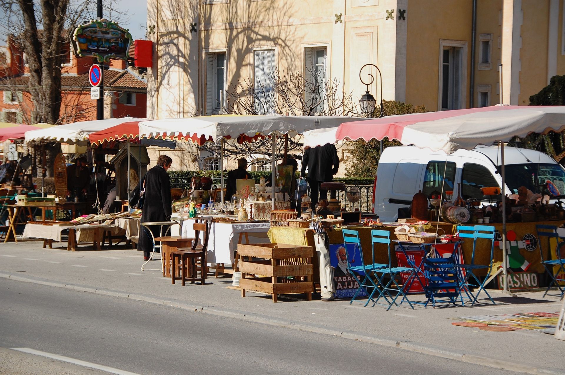 Un marché d'antiquités ensoleillé en plein air, avec des étals sous des auvents, présentant meubles et objets le long d'une rue.