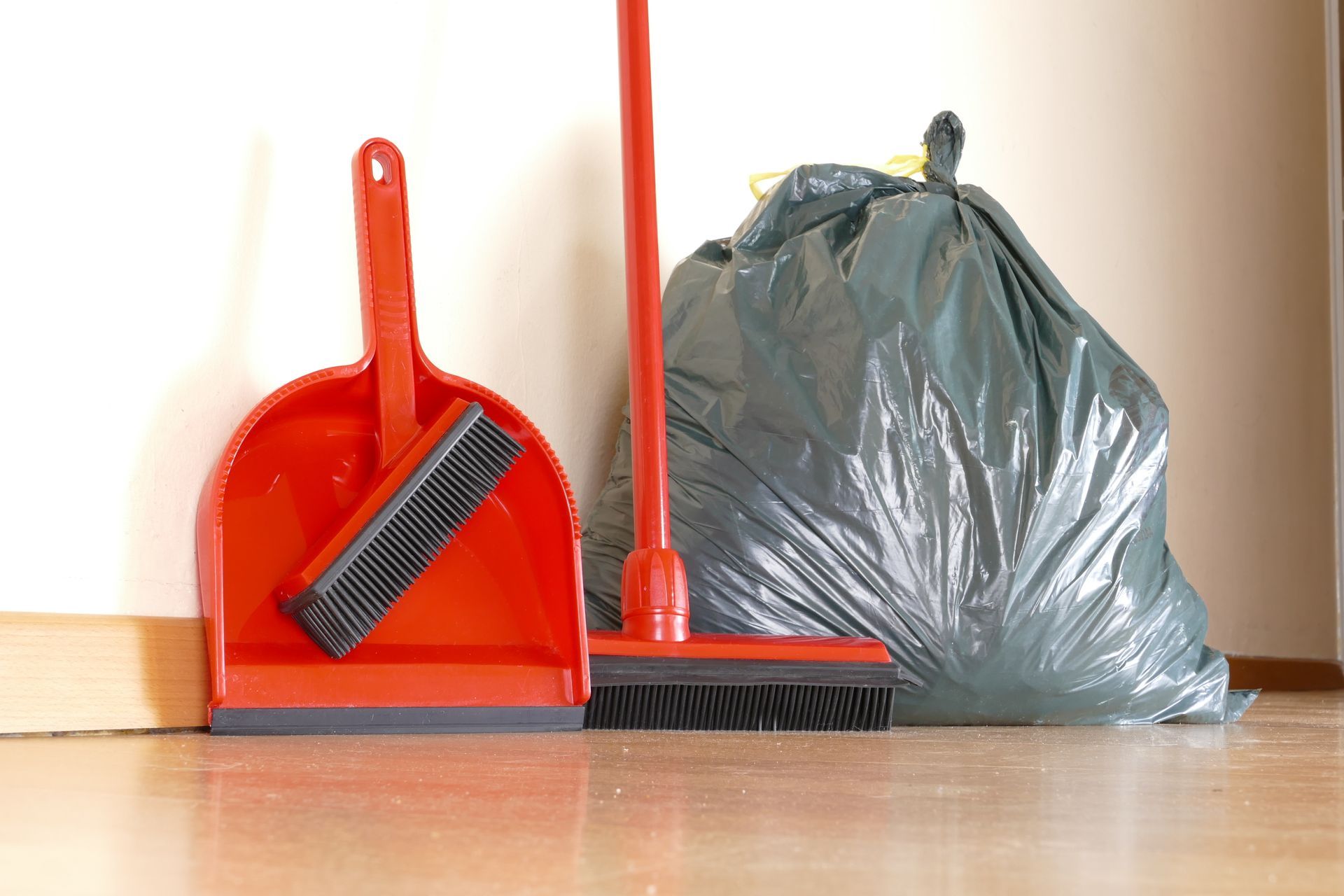 Une pelle à poussière avec une balayette, un balai et un sac-poubelle sont posés sur un plancher en bois.