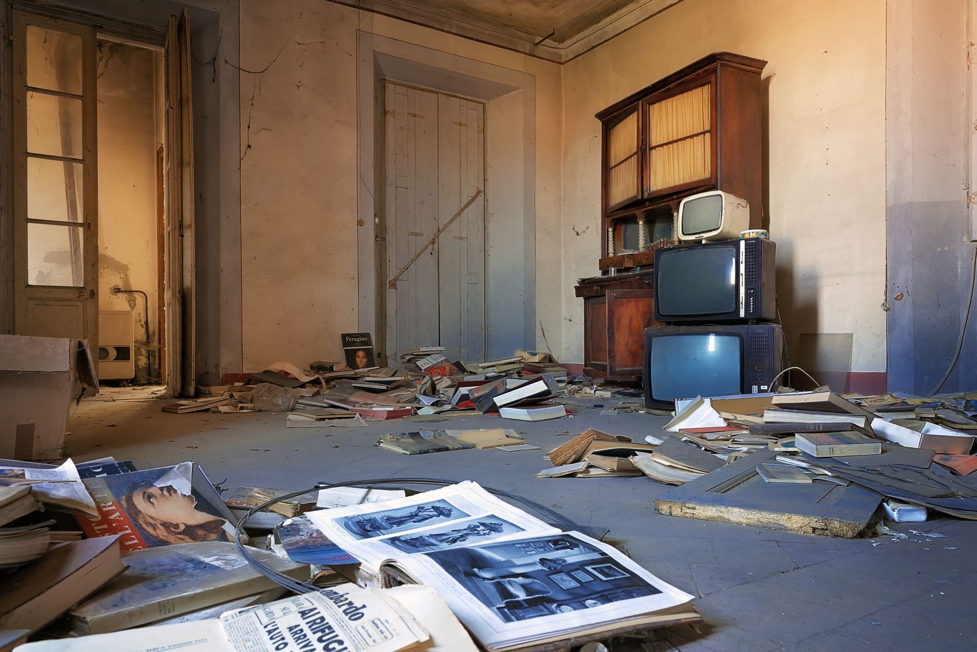 Une pièce abandonnée et poussiéreuse, avec des papiers éparpillés, de vieux livres et une pile de trois téléviseurs vintage à côté d'un buffet en bois.