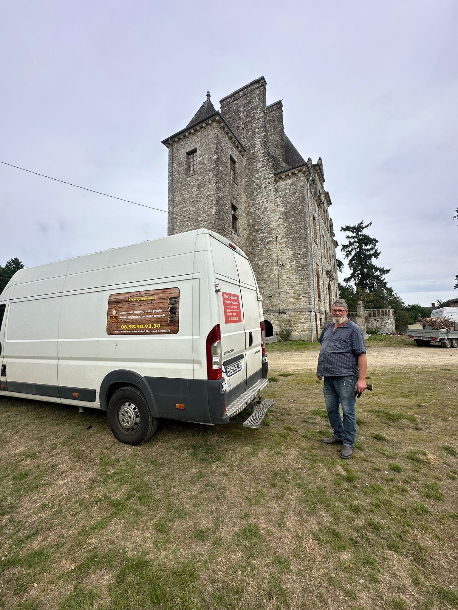 Une personne se tient à côté d'une camionnette garée sur un terrain herbeux devant un grand château en pierre de style médiéval.