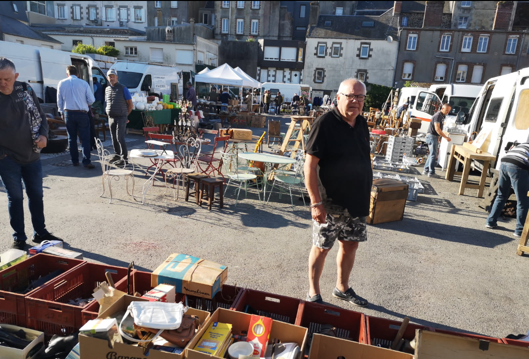 Scène de marché aux puces en plein air, en journée, avec des vendeurs proposant des antiquités, de la verrerie et des objets divers.