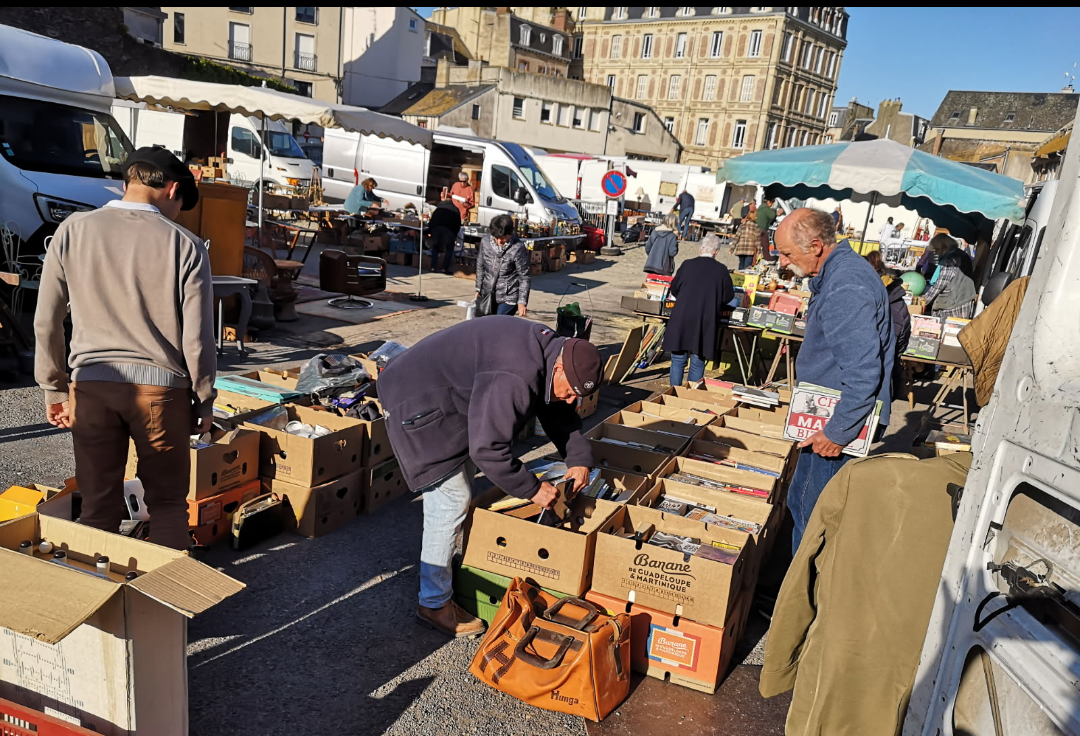 Des gens examinent des cartons de marchandises sur un marché en plein air, par une journée ensoleillée, devant des bâtiments.