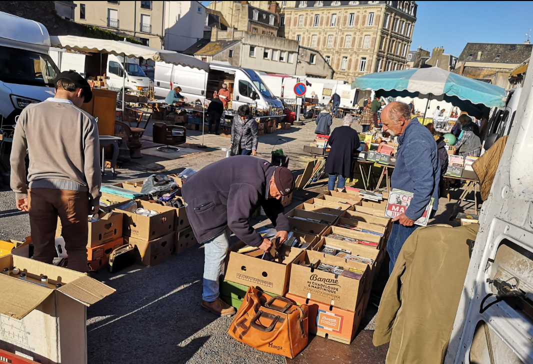 Des passants examinent des cartons d'objets à vendre sur un marché aux puces en plein air, par une journée ensoleillée sur une place de ville.