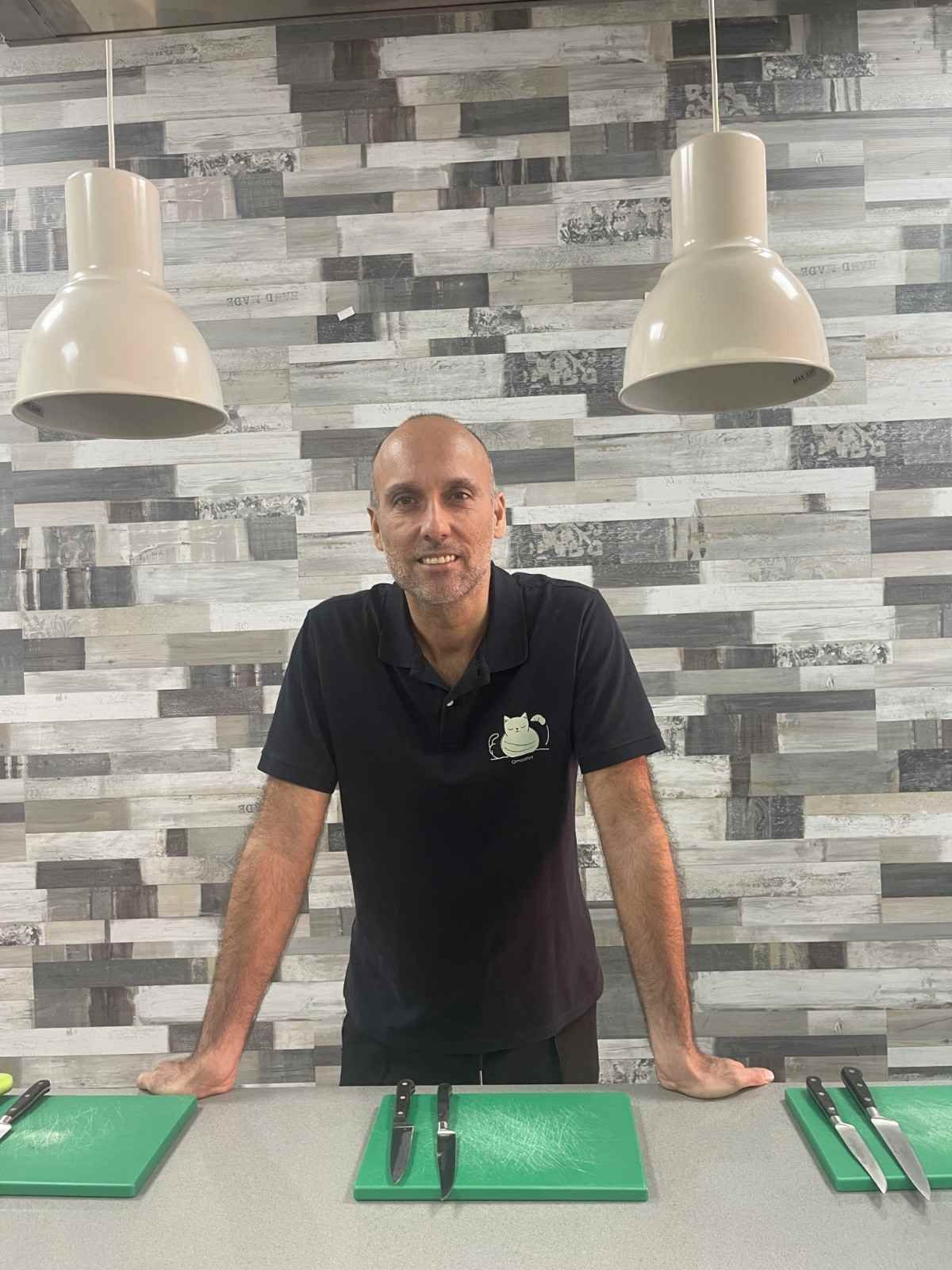 Hombre de pie detrás de una encimera en una cocina con tablas de cortar, utensilios y lámparas colgantes encima
