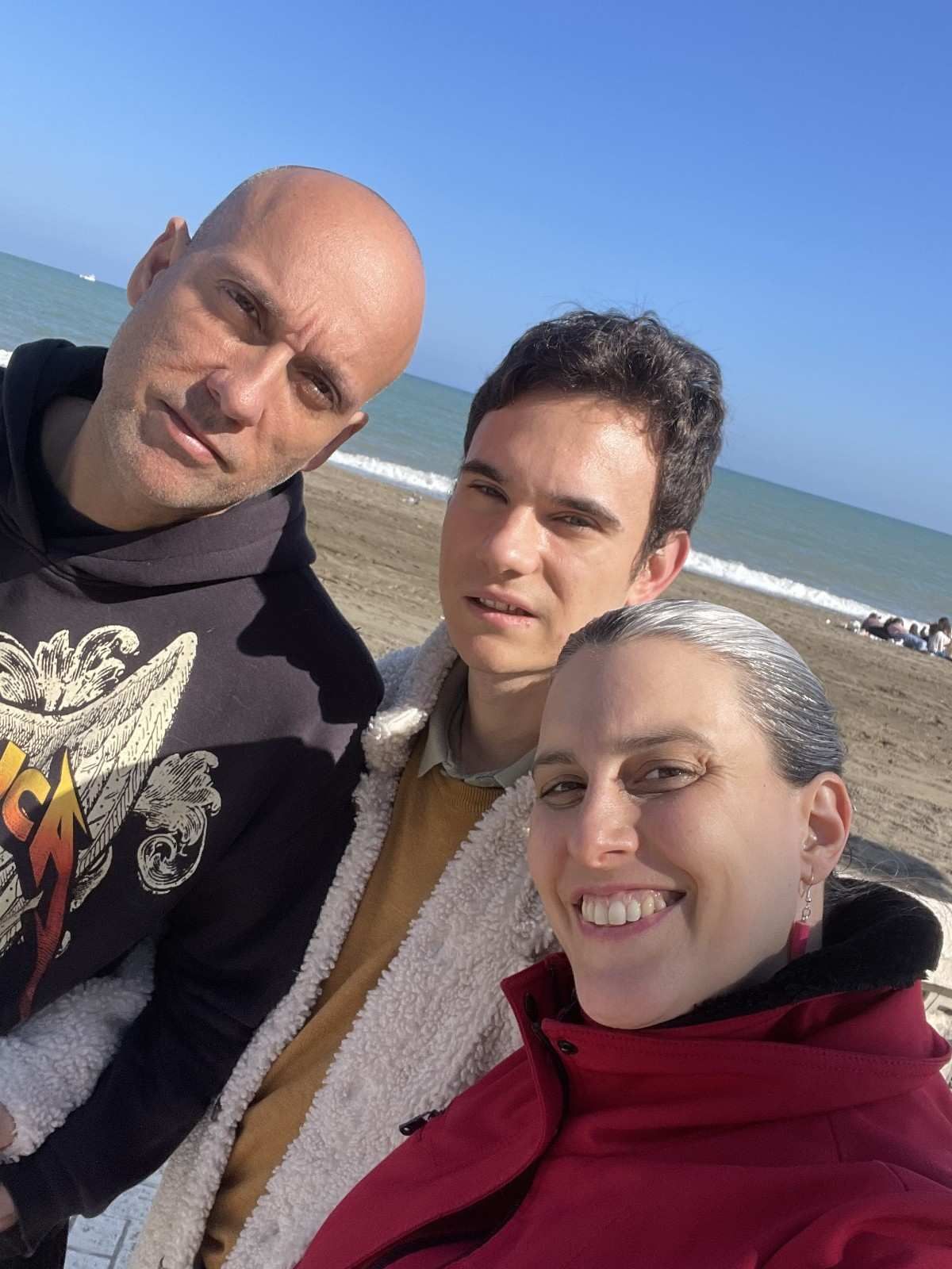 Tres personas sonríen para un selfi en una playa, con el océano y el cielo azul de fondo