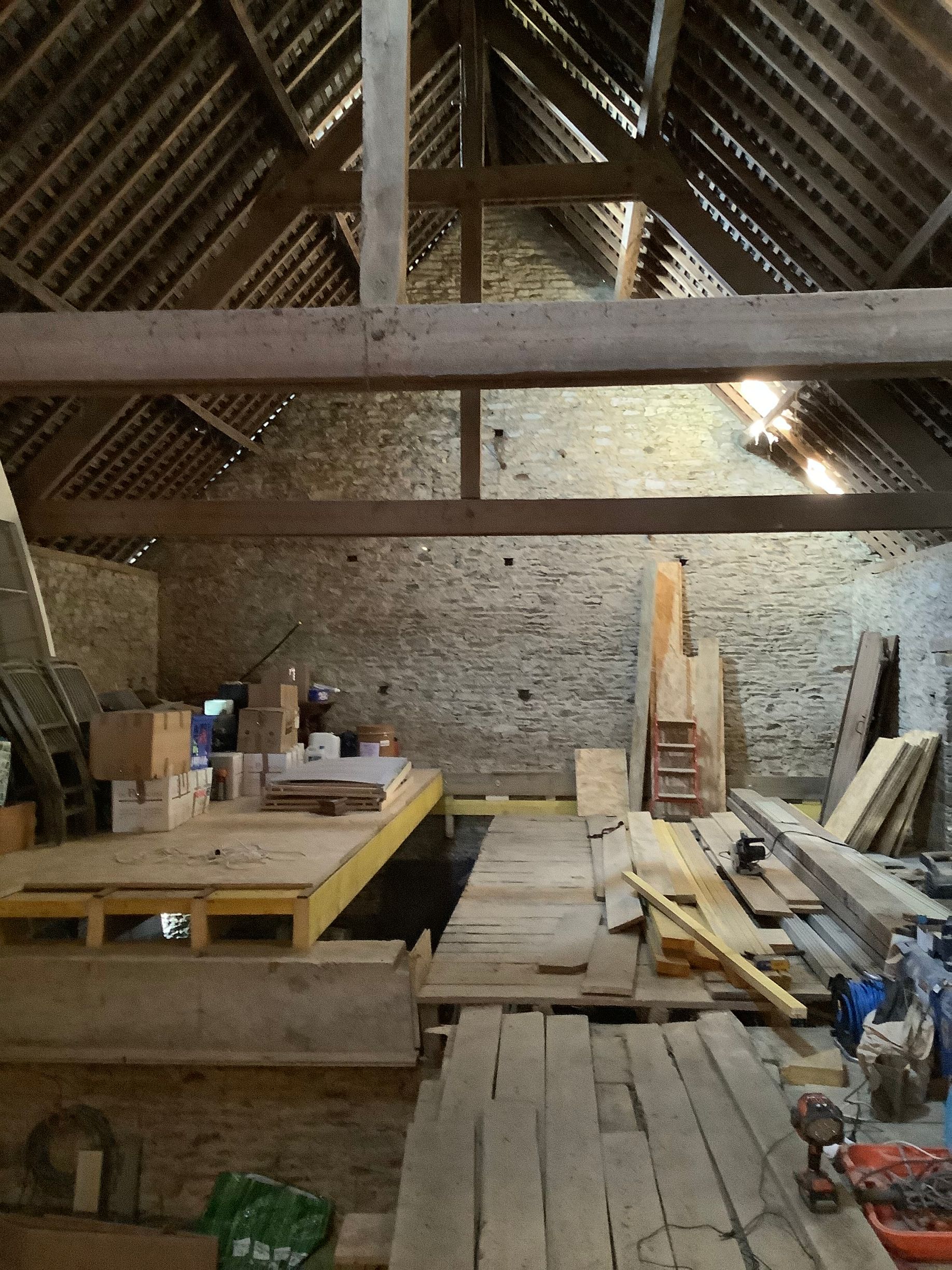 Un grenier en cours de rénovation avec des poutres apparentes, des murs en pierre, des piles de bois et des plateformes en bois.