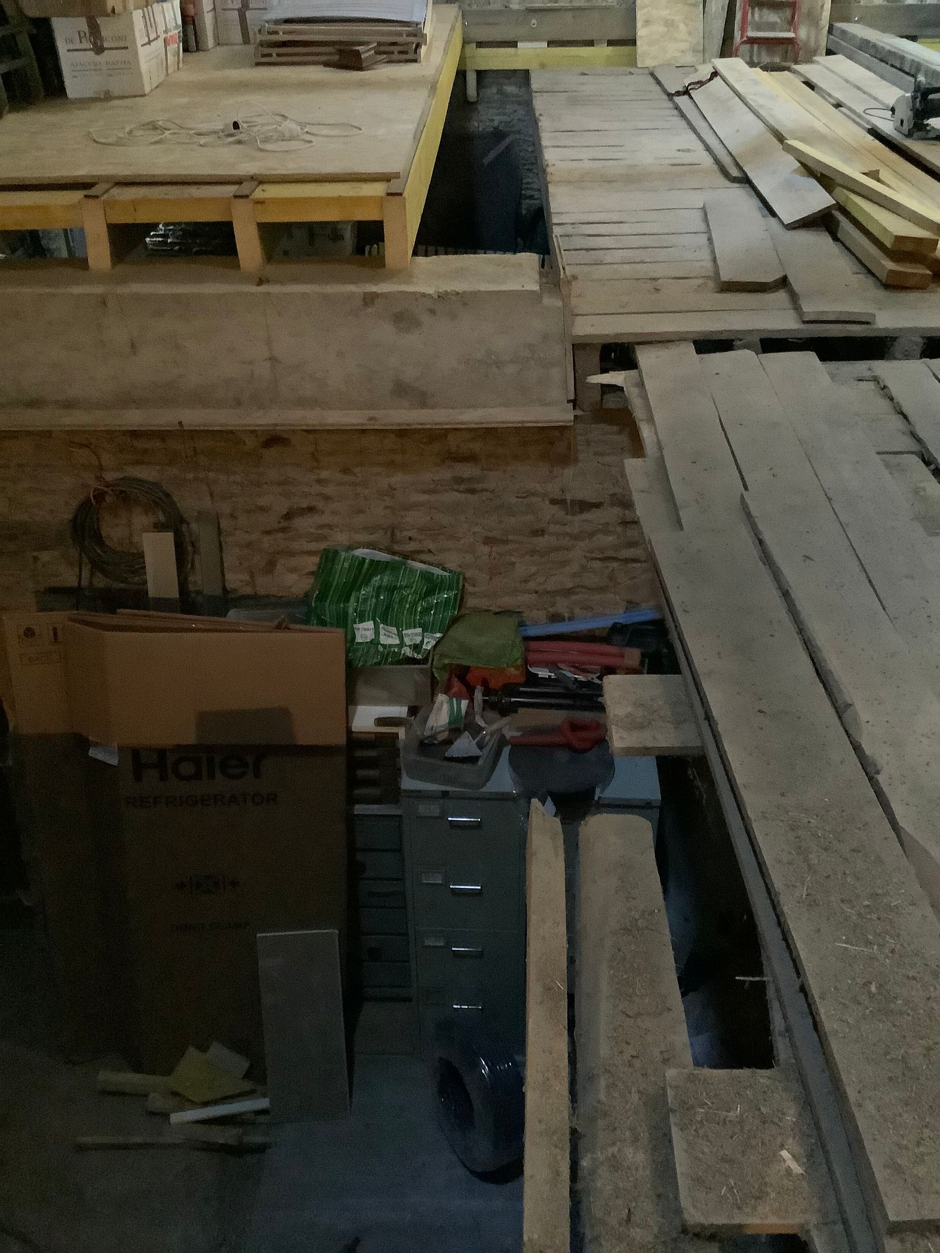 Un sous-sol ou un espace de rangement encombré, avec des plateformes en bois, des piles de bois de construction, des boîtes en carton.