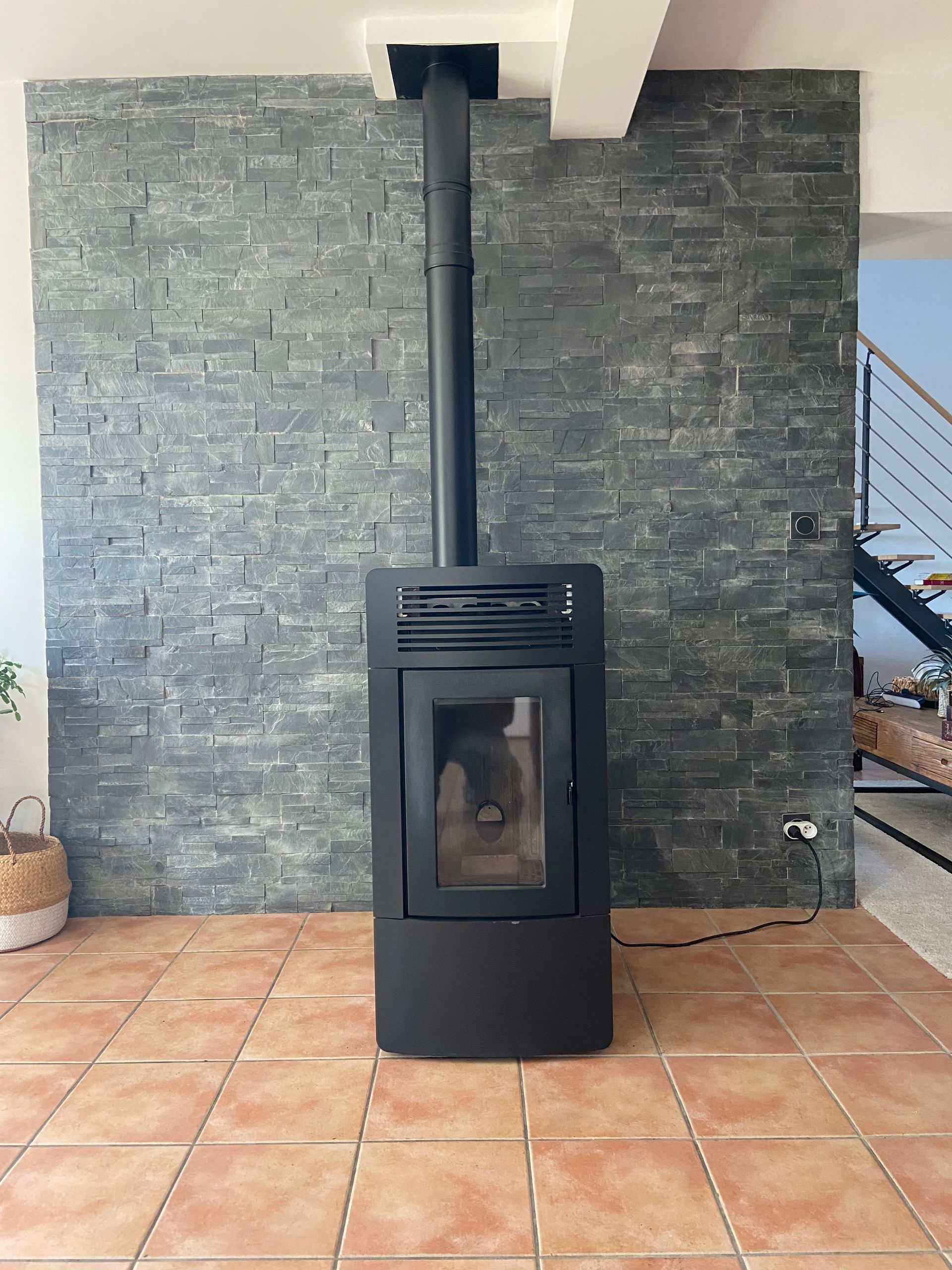 Un poêle à granulés noirs est posé sur des carreaux de terre cuite devant un mur de pierre gris foncé avec un conduit d'évacuation vertical.