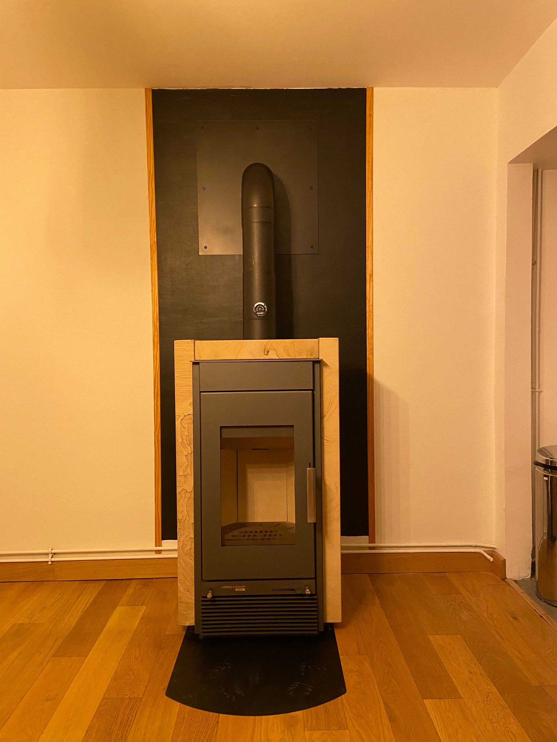 Un poêle à bois, encadré d'un cadre en bois clair, contre un panneau mural noir muni d'un conduit d'évacuation.